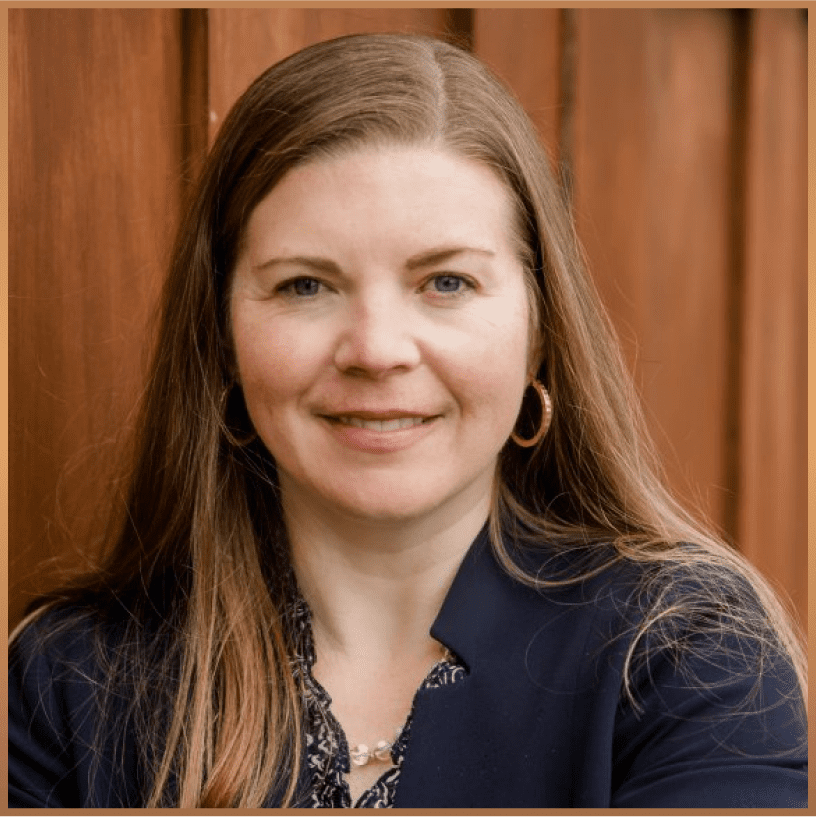 A woman with long brown hair, wearing a navy blazer, patterned blouse, hoop earrings, and a necklace, smiles in front of a wooden background.
