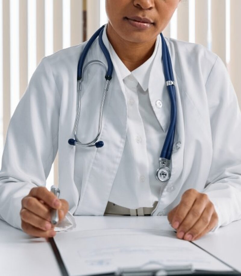 Person in a white medical coat with a stethoscope around their neck sits at a desk, holding a pen and writing on a clipboard.