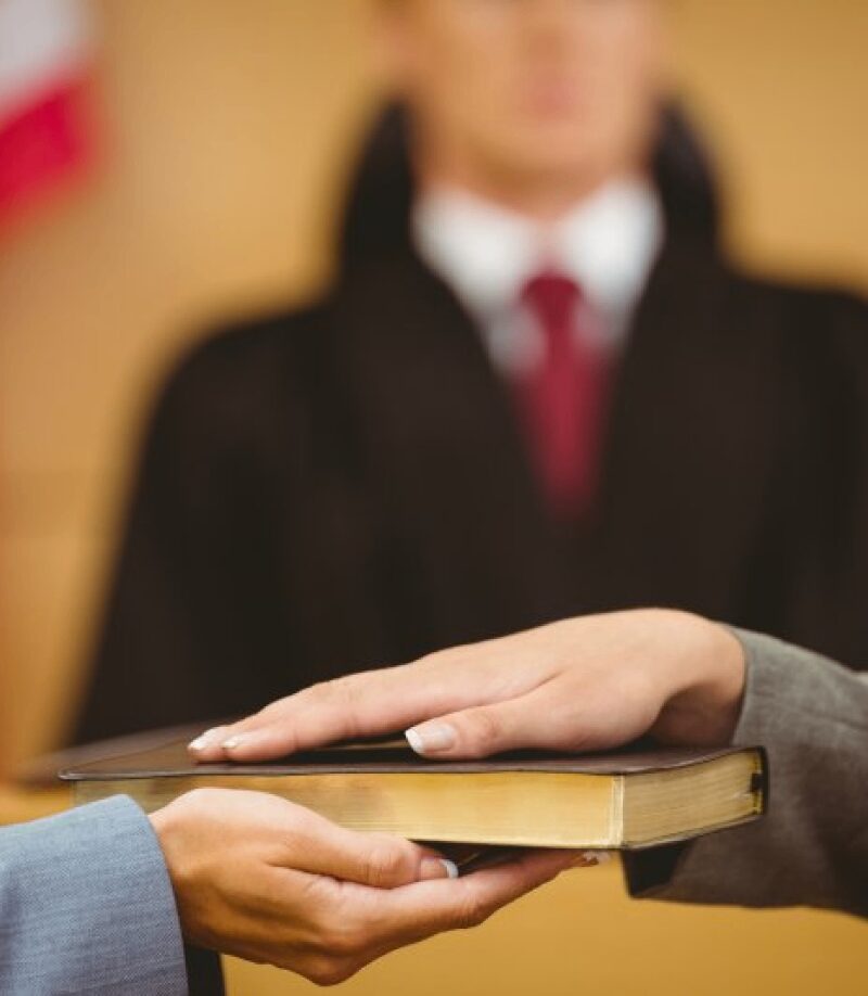 A person places their hand on a book, likely to take an oath, with a judge in robes and an American flag blurred in the background.