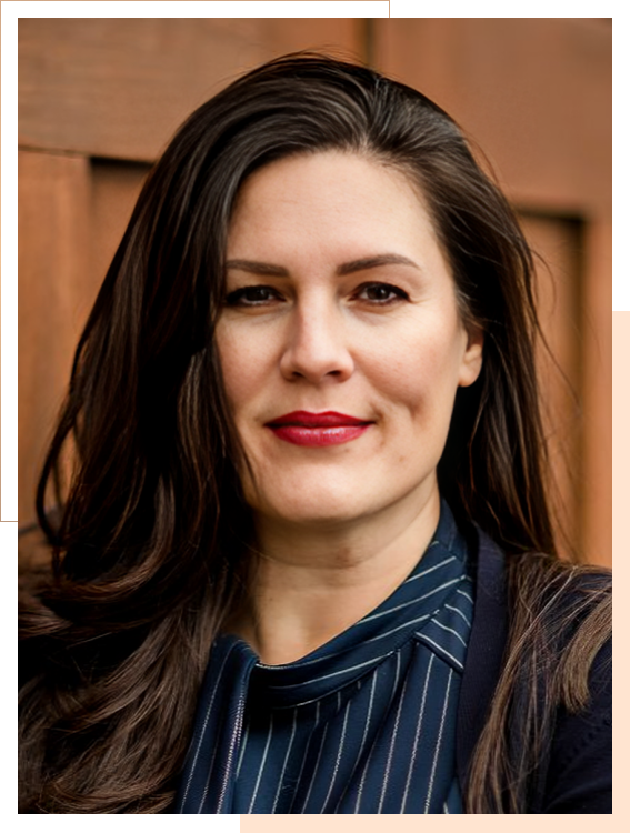 A woman with long dark hair, wearing a dark striped blouse and blazer, stands in front of a wooden background, looking at the camera.