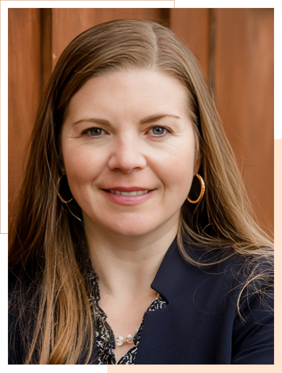 A woman with long light brown hair, wearing gold hoop earrings, a dark blazer, and a patterned blouse, stands in front of a wooden background, looking at the camera.