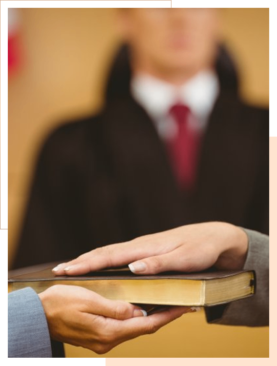A person places their hand on a book, likely taking an oath in court, with a judge in a robe visible in the background.